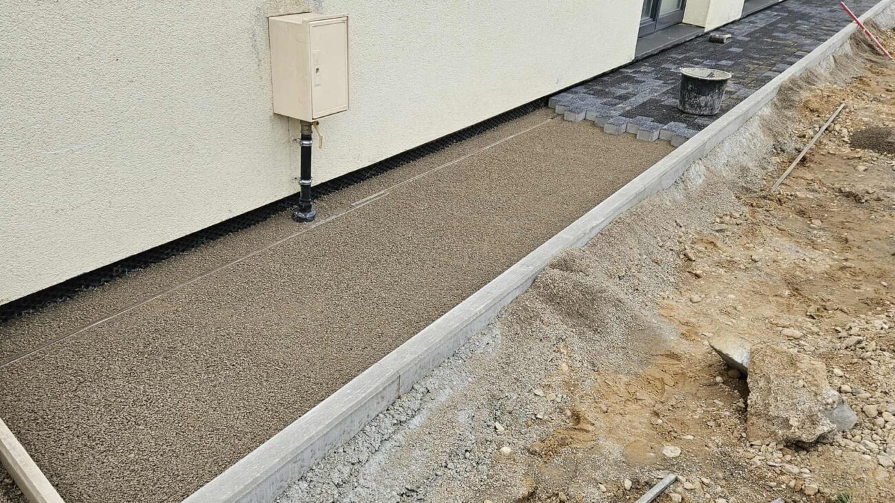 Trinkelių pagrindo paruošimas prie namo: išlygintas atsijų sluoksnis tarp bortelių